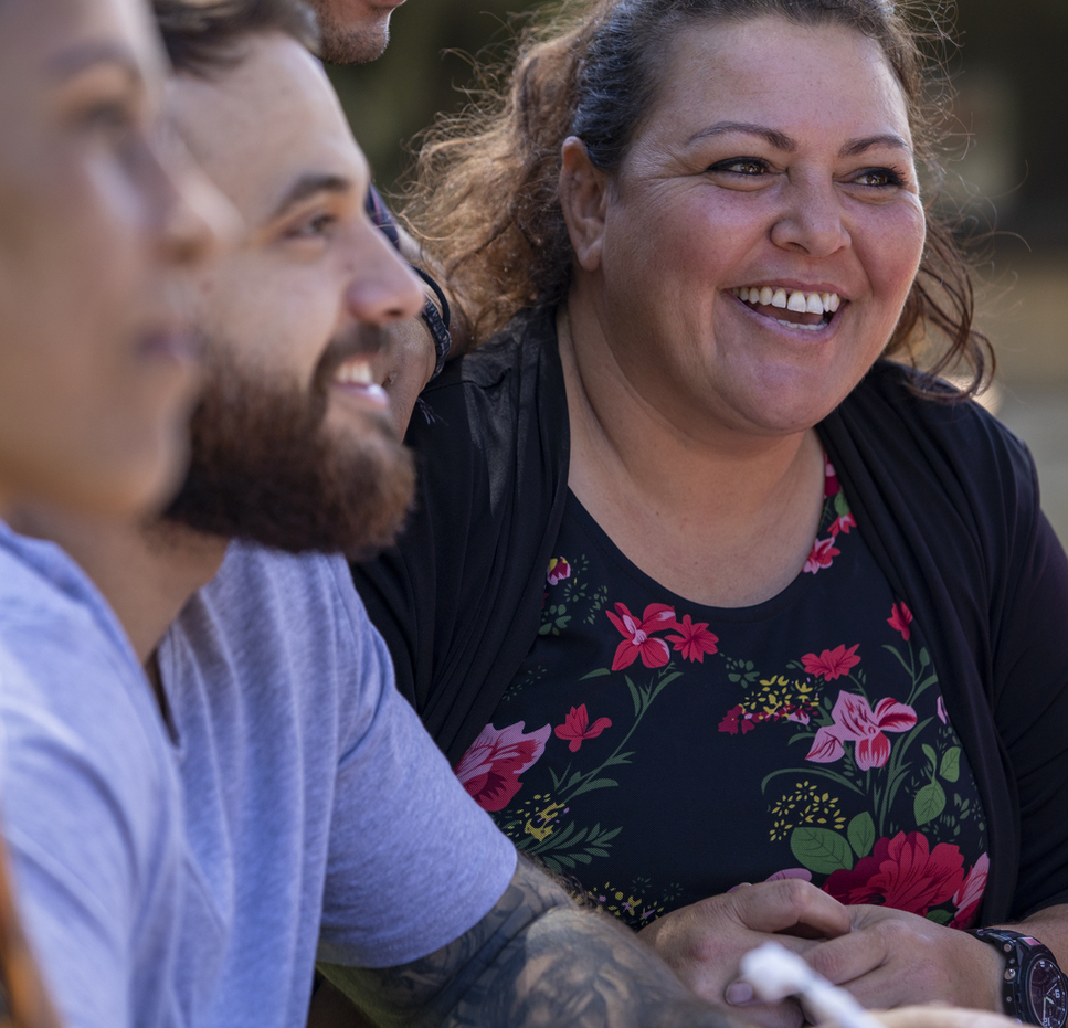 Close-up of a smiling woman in a floral dress talking with others during an outdoor group meeting.