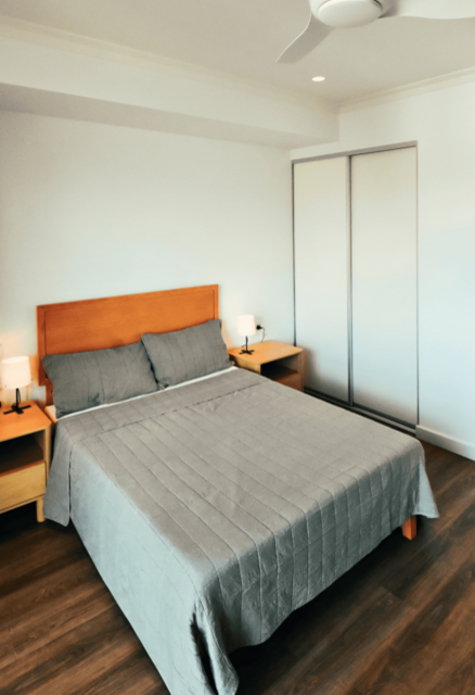 A modern bedroom with a neatly made bed, bedside tables with lamps, a ceiling fan, and built‑in wardrobe doors.