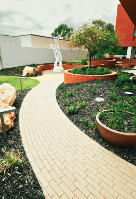 A curved paved pathway leading through a landscaped garden with plants, raised garden beds, large rocks, and an outdoor sculpture.