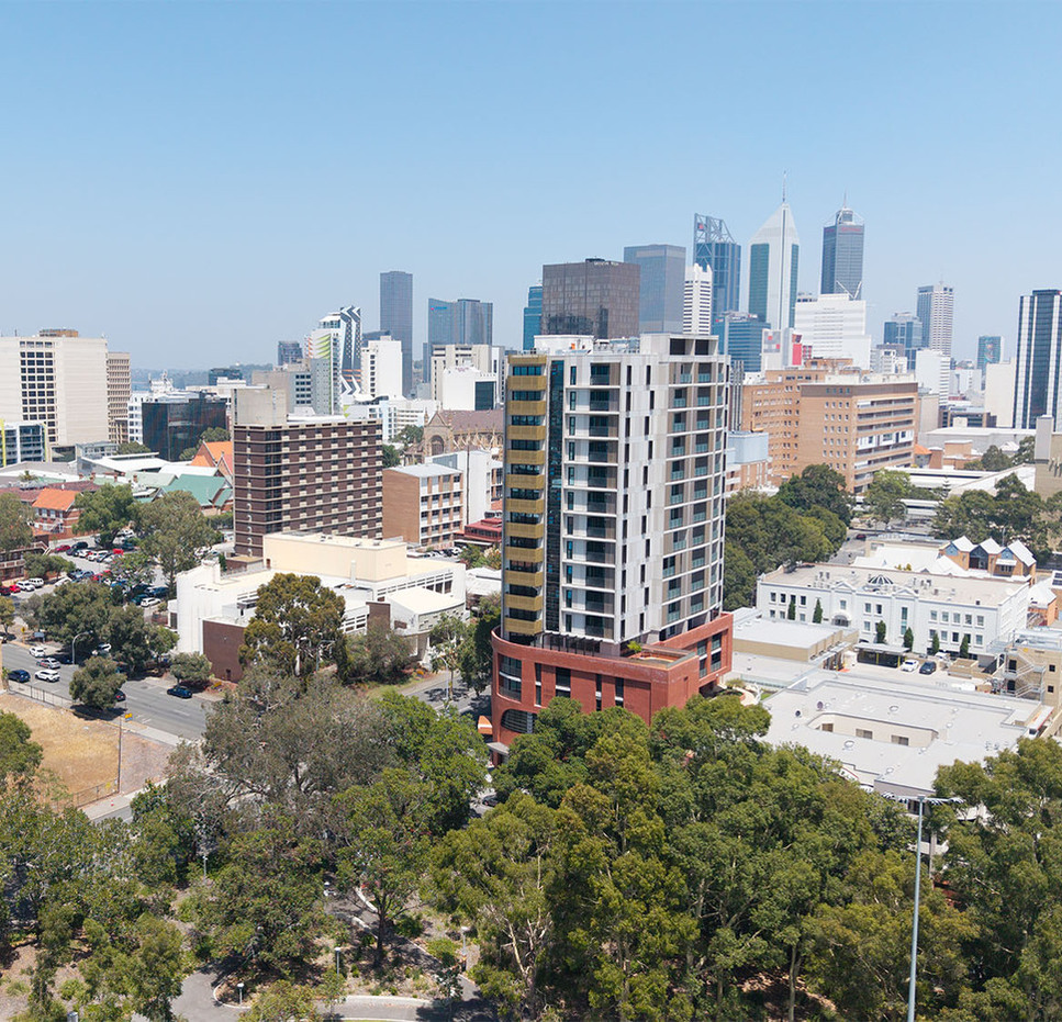 Aerial view of the Common Ground East Perth apartment building, with surrounding homes, trees and the Perth city skyline in the background.