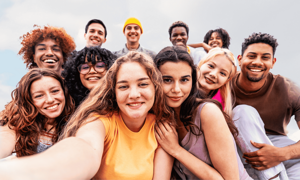 A group of young people gather closely outdoors to take a group selfie.