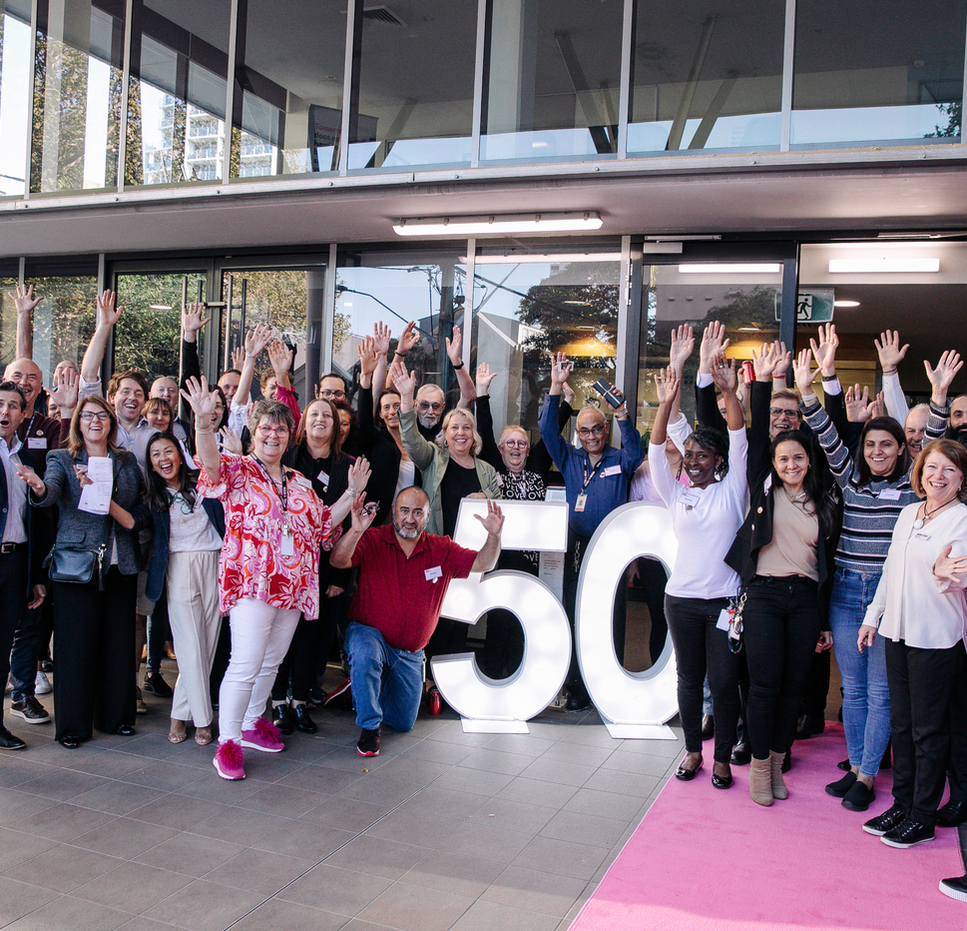 A large, diverse group of Mission Australia staff members stand smiling and waving around a bright, illuminated “50” sign outside a building, celebrating 50 years of MAC Surry hills together.