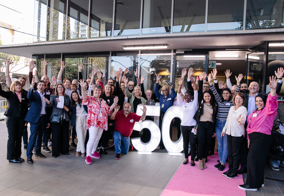 A large, diverse group of Mission Australia staff members stand smiling and waving around a bright, illuminated “50” sign outside a building, celebrating 50 years of MAC Surry hills together.