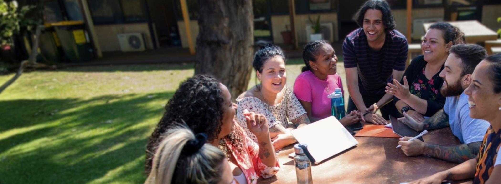A group of people sit around an outdoor table in discussion, with notebooks, devices, and personal items visible.