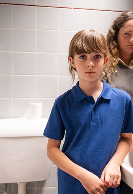 Mother and child in bathroom with tiled walls and a mirror. One stands near the sink holding a cloth; the other is in the foreground with hands clasped.