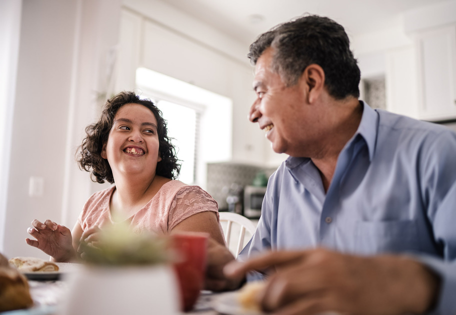 A daughter who appears to live with disability and her father sitting in the kitchen, smiling at each other while having breakfast.