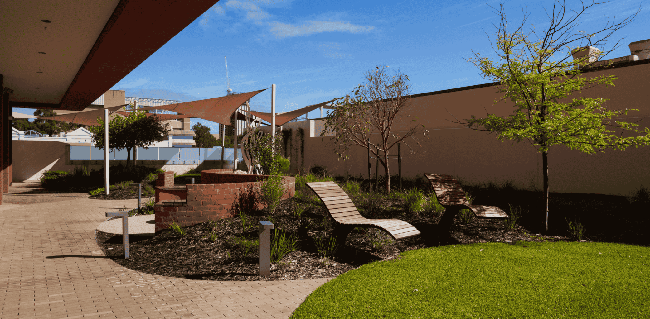 A landscaped courtyard at Common Ground East Perth with paved paths, seating, plants, and shade structures under a blue sky. 