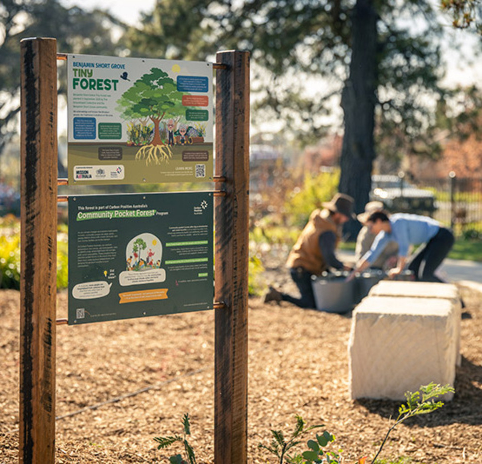 Two community information signs for the Benjamin Short Grove Tiny Forest and Community Pocket Forest project stand in a garden bed at Mission Australia’s aged care facility in Orange, while two people work together planting seedlings in the background among trees and garden pathways.