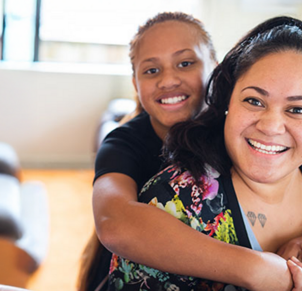 Sally smiling with a young girl hugging her from behind, both at home, featured in Mission Australia's Summer Impact Report 2025.