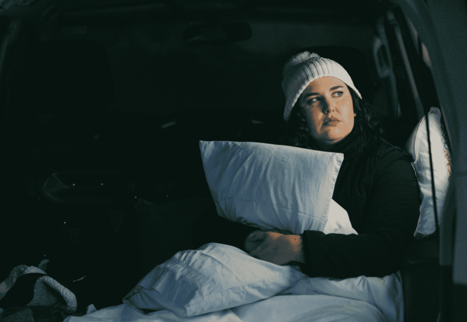 Person sitting inside the back of a vehicle holding a pillow with blankets and bedding around them.