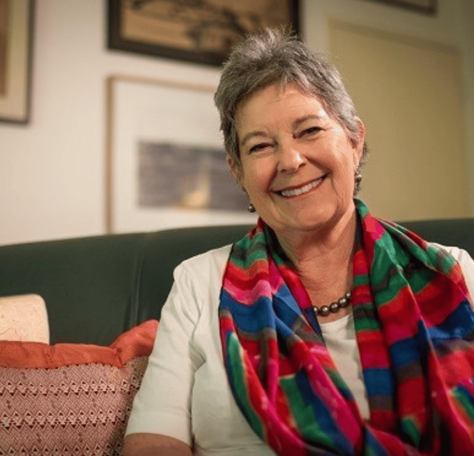 Sue Boyd sitting on a couch with colourful striped scarf and framed artwork on the wall behind.