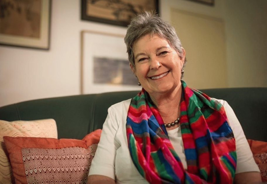 Sue Boyd sitting on a couch with colourful striped scarf and framed artwork on the wall behind.