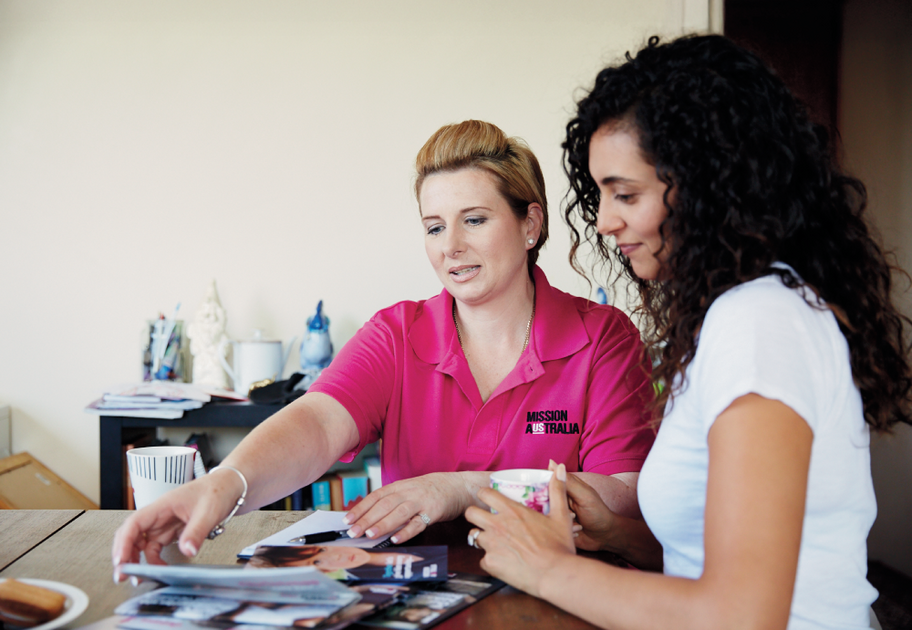 A Mission Australia case worker speaks to a woman with curly brown hair as they look through service brochures on a dining table.