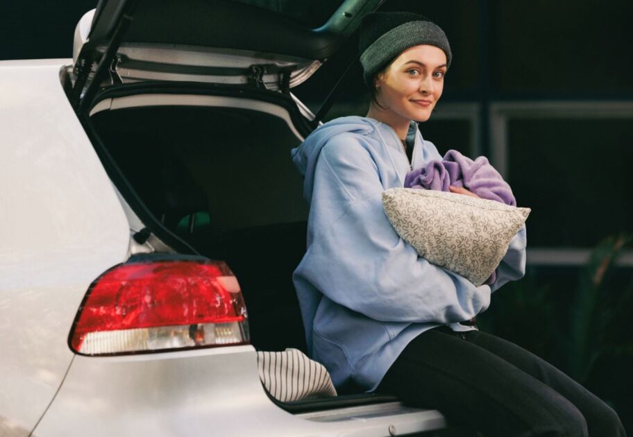 A person sits at the open back of a car holding a pillow and blanket, with the vehicle parked outdoors.