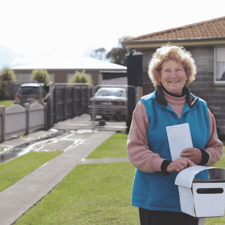 Smiling older woman standing outside in front of her home, holding mail by a white letterbox, with houses and a driveway in the background.