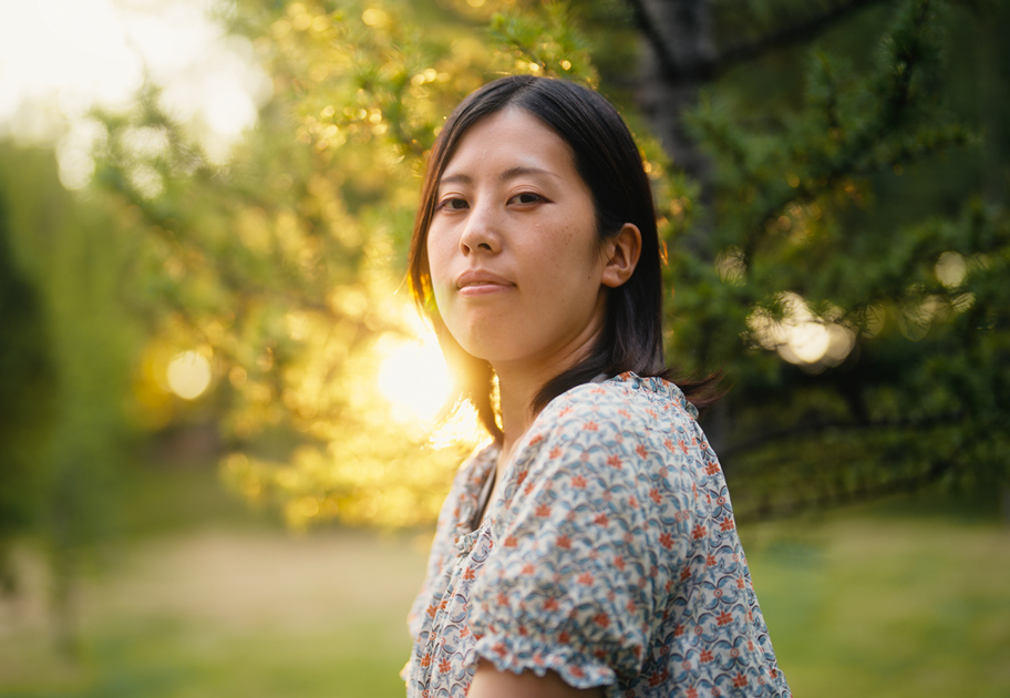 A woman with straight dark hair, wearing a patterned shirt, looks at the camera while standing outdoors in a sunlit, green park with trees and soft sunlight in the background.
