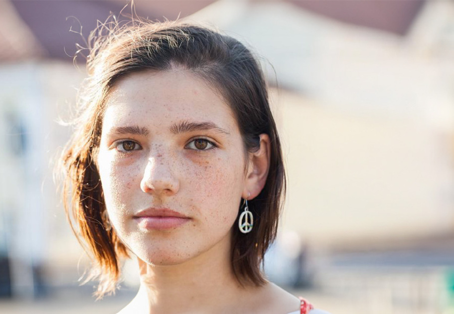 A young person standing outdoors with short hair, wearing earrings, and a light-colored top, with blurred buildings in the background.