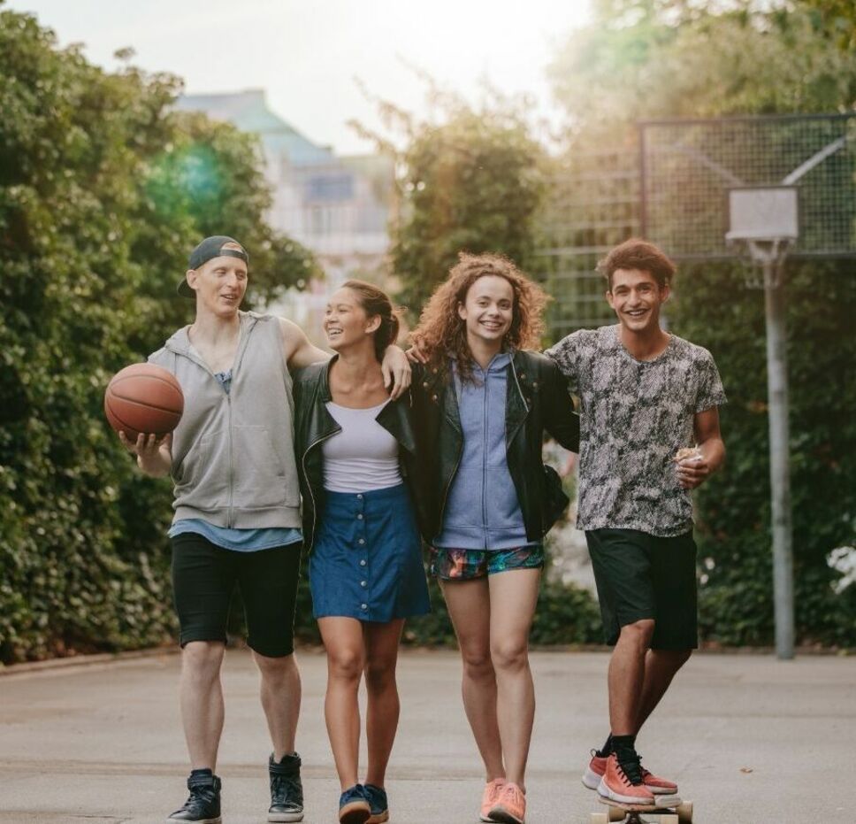 Four smiling young people walking together outdoors, holding a basketball and skateboard.