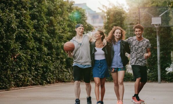 Four smiling young people walking together outdoors, holding a basketball and skateboard.